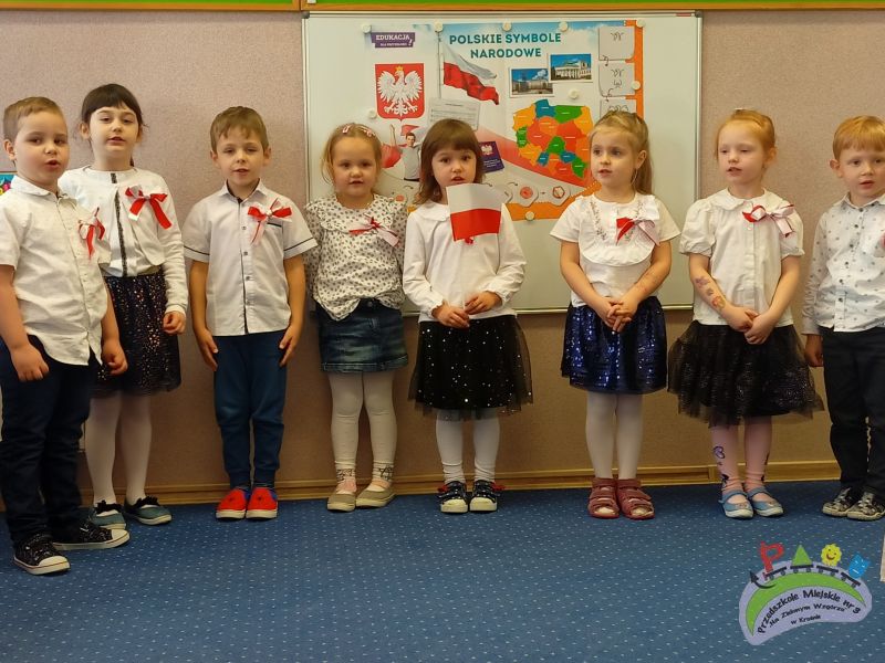 Ośmioro dzieci stoi pod białą tablicą z plakatem „Polskie Symbole Narodowe”. Dzieci ubrane są w biało-granatowe stroje i biało-czerwone kotyliony. Dziewczynka stojąca po środku trzyma flagę polski