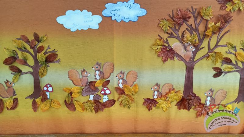 Siedem rudych wiewiórek siedzących na jesiennych liściach, drzewie, grzybach. Liście są koloru żółtego i pomarańczowego, znajdują się na trzech drzewach i pod nimi. Na górze widnieją dwie chmury: niebieska i biała