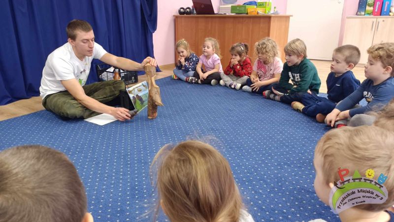 Pan leśnik siedzi na granatowym dywanie i pokazuje dzieciom małą figurkę bobra i prawdziwy kawałek drewna obgryzionego przez bobra, a obok ustawione jest zdjęcie bobra. Dzieci siedzą wokół i obserwują zajęcia