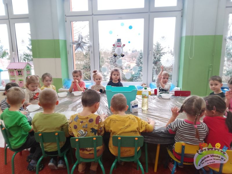 Na tle zimowego okna, na którym widać śnieżynki i bałwanka, przy stole siedzi grupa dzieci, gotowych do zabaw sensorycznych. Dzieci wykonają sztuczny śnieg, ulepią śniegowe kule i będą uczestniczyły w zabawach muzyczno - ruchowych.
