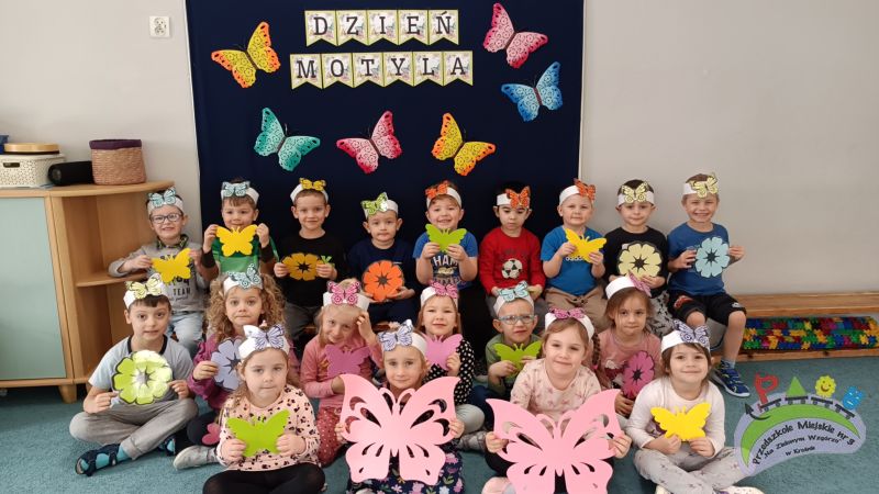 Na tle dekoracji z kolorowymi motylkami, na której widnieje napis „Dzień Motyla”, siedzi grupa uśmiechniętych dzieci w opaskach – motylkach na głowie. Dzieci trzymają motylki i kwiatki. Przedszkolaki są gotowe do wspólnej zabawy z okazji „Dnia Motyla”.