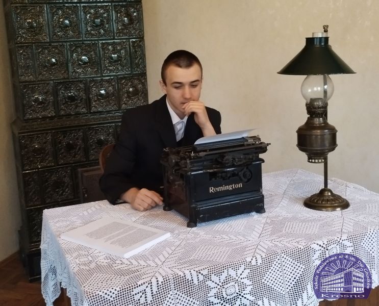 Zdjęcie przedstawia ucznia Igora Barana wcielającego się w rolę Stefana Żeromskiego. Uczeń siedzi przy stole, na którym znajduje się lampa z końca XIX w. oraz maszyna do pisania z początku XX w. Całość dopełnia piękny piec kaflowy
